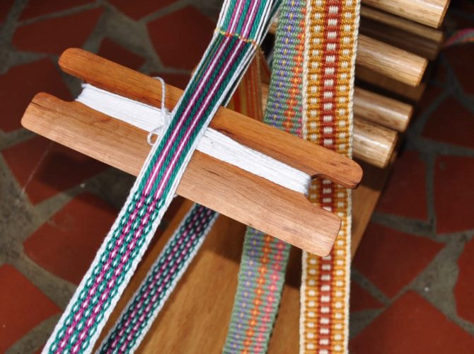 Image of an inkle loom with weaving in progress, and two additional finished inkle bands.